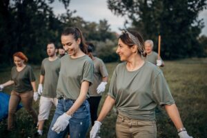 A group of volunteers wearing gloves and green shirts participates in an outdoor cleanup event, smiling as they walk through a grassy area with trees in the background.